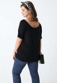 Zwarte top met korte mouwen en geschulpte kanten accenten, gecombineerd met blauwe denim jeans en een kleine zwarte handtas, gezien van achteren.