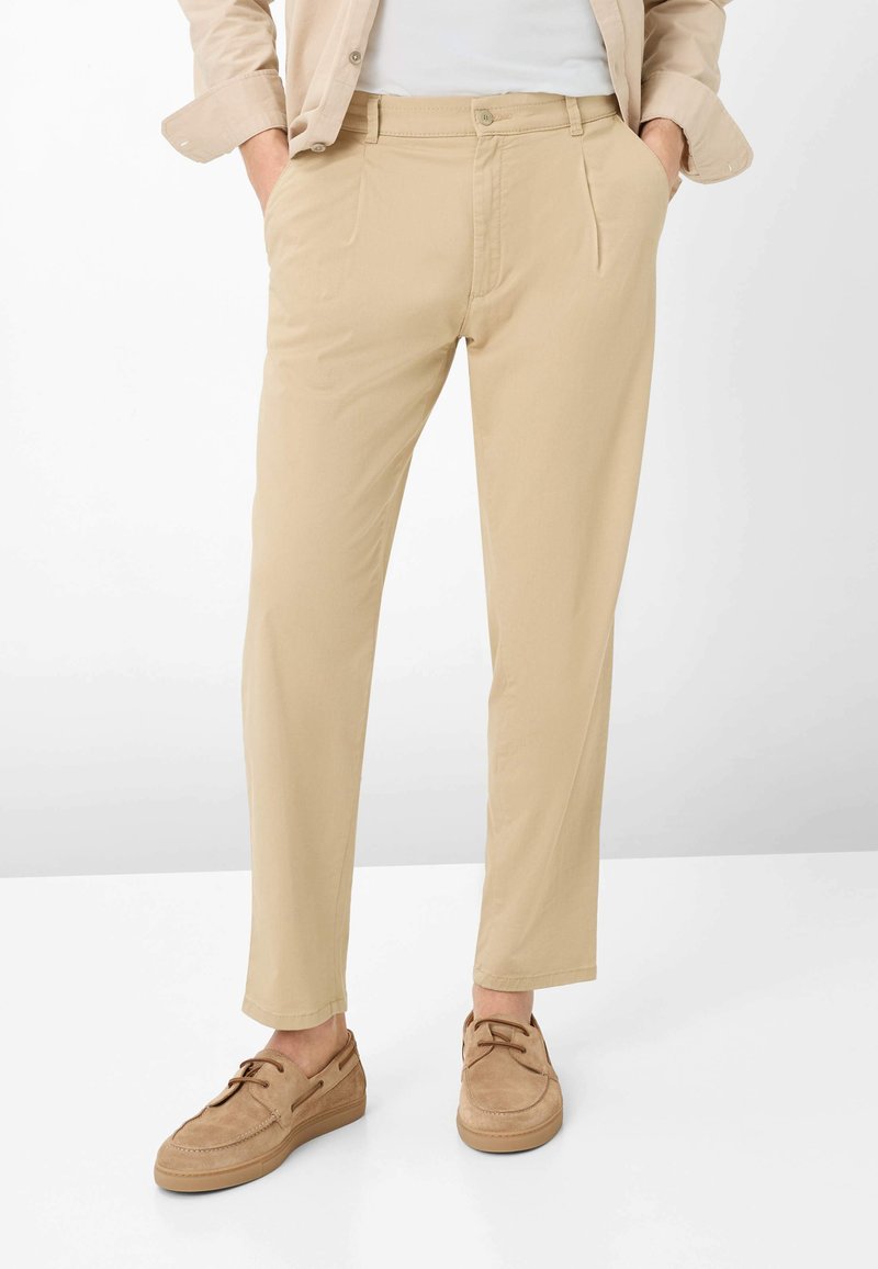 Pantalon beige à jambes droites avec plis devant, porté avec des chaussures en daim marron clair à lacets, une veste beige et une chemise blanche, mains dans les poches.