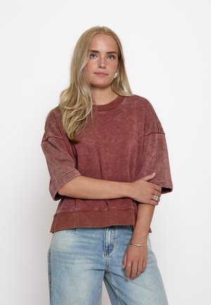 Frau mit langen blonden Haaren, die ein lockeres, ausgebleichtes rotes kurzärmeliges Oberteil und hellblaue Jeans trägt, steht mit verschränkten Armen vor weißem Hintergrund.