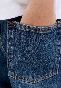 Sötétkék mosott farmernadrág látható textúrával. A hátsó zseb látható, amely kontrasztos varrással és szabványos téglalap alakú formával rendelkezik.