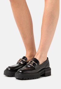 Zwarte leren instappers met een brede band en een ronde metalen accent, met een chunky platform zool met gestructureerde groeven.