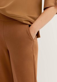 Bronzefarbenes Oberteil mit lässiger Passform, das eine glatte Textur und weite Ärmel aufweist. Braune Hose mit Seitentaschen und einer weichen Oberfläche.