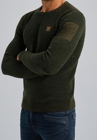 Maglione verde lavorato a maglia con colletto a girocollo, tessuto strutturato e dettagli con toppe sulle spalle. Abbinato a jeans in denim blu.