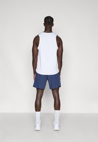 Άνδρας με πλεξούδες που φοράει λευκό άνοιχτο αμάνικο μπλουζάκι, ναυτικά μπλε σορτς, λευκές κάλτσες και λευκά αθλητικά παπούτσια, στέκεται με την πλάτη προς την κάμερα.