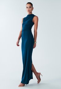 Turquoise mouwloze maxi-jurk met een hoge hals, een geplooide taille, een zijsplit en een gladde, rekbare stof; gecombineerd met nudekleurige sandalen met hak.