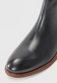 Bocanci din piele la gleznă, negri, cu o textură netedă, vârf ascuțit și un accent maro pe marginea tălpii. Design minimalist cu linii elegante.