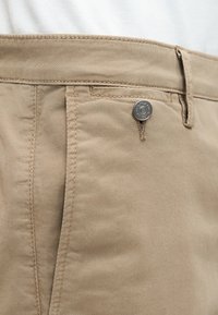 Κοντινό πλάνο σε τσέπη μπεζ παντελονιού με μεταλλικό κουμπί και θηλιά για ζώνη, που δείχνει την υφή του υφάσματος και τις λεπτομέρειες του ραψίματος.