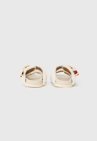 Sandales sans dos à fourrure blanche, avec une semelle beige lisse et une petite étiquette multicolore sur la sangle.