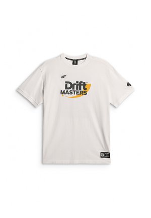 Młody mężczyzna o jasnych włosach, ubrany w białą koszulkę Drift Masters i czarne spodnie, stoi na jednolitym szarym tle.
