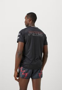 Schwarzes Sport T-Shirt mit kurzen Ärmeln, auf dem Rücken mit rotem Logo. Getragen mit schwarzen Shorts, die am Saum mit rotem Zickzackmuster verziert sind.