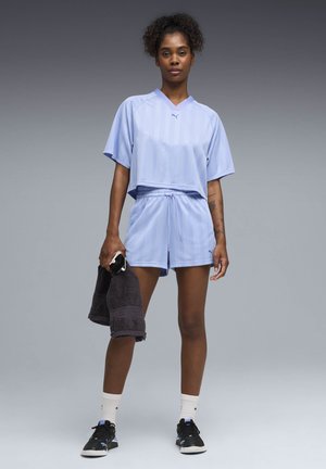 Donna con pantaloncini sportivi e top viola chiaro, che tiene un asciugamano nero e uno smartphone, indossando calzini bianchi e scarpe da ginnastica nere, in piedi su sfondo grigio.