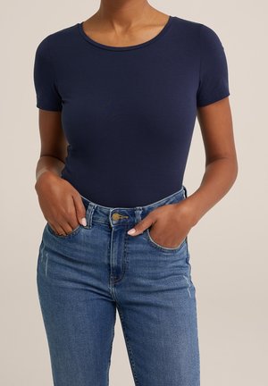 Vrouw draagt een nauwsluitend, marineblauw shirt met korte mouwen, ingestopt in een hoge taille blauwe spijkerbroek, met haar handen in de voorzakken.