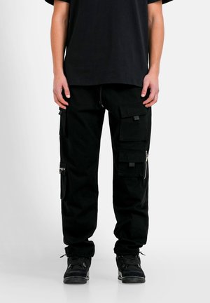 Personne portant un pantalon cargo noir avec plusieurs poches et des baskets noires, debout devant un fond clair uni.