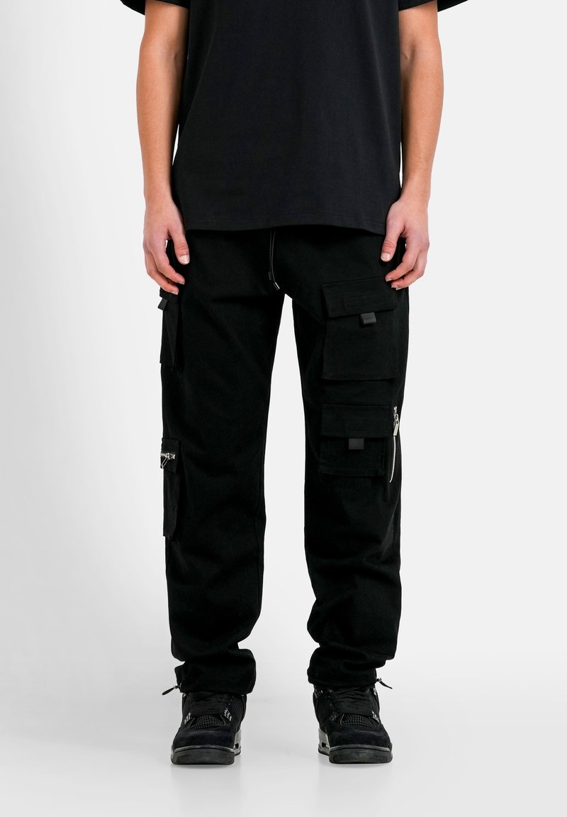 Personne portant un pantalon cargo noir avec plusieurs poches et des baskets noires, debout devant un fond clair uni.
