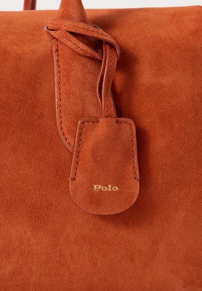Oranžs zamšs roku soma ar teksturētu virsmu, ādas birku un zelta "Polo" logotipu, kas iepresēts birkā.
