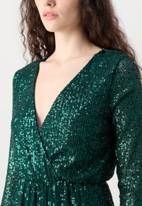 Abito con paillettes in un profondo verde smeraldo. Presenta una scollatura a V, maniche lunghe e un tessuto testurizzato con paillettes luccicanti su tutta la superficie.