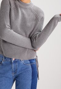 Grijze geribbelde trui met een hoge hals en lange mouwen, voorzien van een strikdetail rondom de taille. In combinatie met hoge taille blauwe jeans.