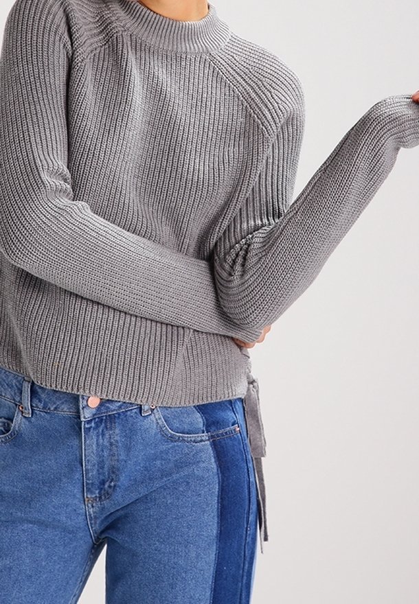 Grijze geribbelde trui met een hoge hals en lange mouwen, voorzien van een strikdetail rondom de taille. In combinatie met hoge taille blauwe jeans.