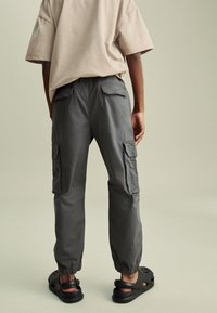 Pantaloni cargo grigi con polsini elasticizzati e molteplici tasche laterali, abbinati a calzature nere scivolose. Vestibilità casual e texture morbida.