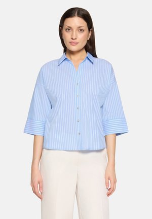 Donna che indossa una camicia azzurro chiaro a righe con maniche larghe a tre quarti e pantaloni bianchi, in piedi davanti a uno sfondo semplice.
