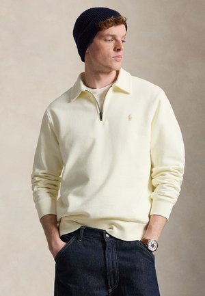 Νέος άνδρας που φοράει μπεζ πουλόβερ με μισό φερμουάρ, σκούρα τζιν, ναυτικό σκουφί και καφέ δερμάτινο ρολόι, στέκεται με τα χέρια στις τσέπες.