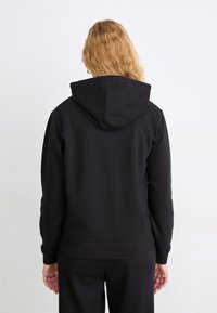 Sudadera negra con capucha, hecha de tejido suave. Presenta mangas largas, puños y dobladillo de canalé, y una textura suave. Mostrada desde la parte de atrás.