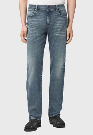 Frontview af person iført falmede blå straight-leg jeans og sorte ruskindsstøvler, stående mod en ensfarvet baggrund.