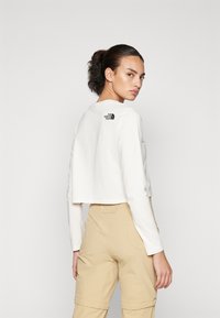 Maglietta bianca a maniche lunghe cropped con scollo rotondo e logo nero sulla schiena, abbinata a pantaloni cargo beige con tasche laterali.