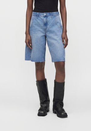 Personne portant un short en denim bleu clair jusqu'aux genoux, un top noir sans manches et des bottes noires mi-mollet, debout devant un fond uni.