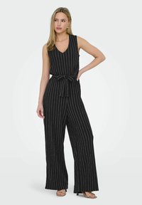 Mono negro con rayas blancas verticales, escote en forma de V y cintura ceñida con cinturón. Presenta piernas anchas y una textura suave. Se lleva con sandalias de tiras.