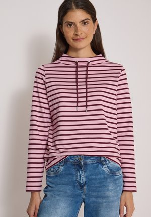Femme aux longs cheveux foncés portant un haut rayé rose et rouge foncé à manches longues et un jean bleu, debout devant un fond uni.