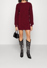 Donna che indossa un vestito a maglia a trecce color borgogna, tiene in mano una borsa nera e indossa stivali da cowboy neri con ricami bianchi.
