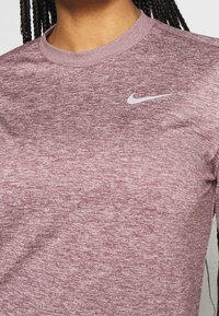 Maglietta sportiva a maniche lunghe color marrone scuro con tessuto strutturato. Presenta un piccolo logo Nike bianco sulla parte superiore sinistra del petto.