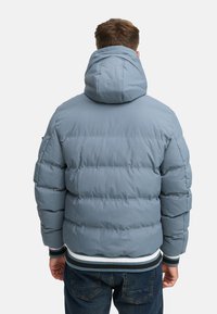 Chaqueta de plumón azul claro con capucha, que presenta costuras horizontales, puños elásticos y un dobladillo acanalado con franjas blancas y azul oscuro.