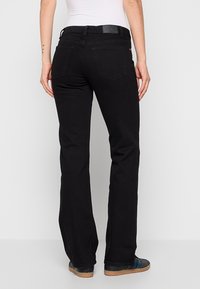 Femme portant un jean noir droit et des baskets noires à rayures bleues, debout, bras détendus le long du corps, dos tourné vers le spectateur.