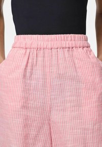 Šviesiai rožinės kelnės su baltomis vertikaliomis juostelėmis, elastinga juosmens juosta ir šoninėmis kišenėmis, pagamintos iš minkštos, tekstūruotos medžiagos.