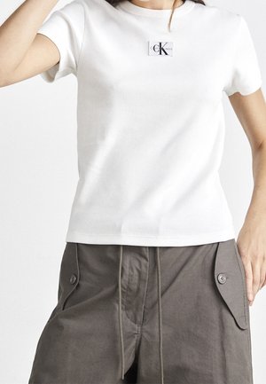 Weißes, geripptes T-Shirt mit kurzen Ärmeln und einem kleinen CK-Logo-Tag auf der Brust. Kombiniert mit grauen Cargo-Shorts mit Seitentaschen.