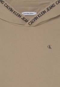 Beige mikina Calvin Klein Jeans s černým nápisem "Calvin Klein Jeans" na okraji kapuce a malým černým logem "cK" na hrudi.