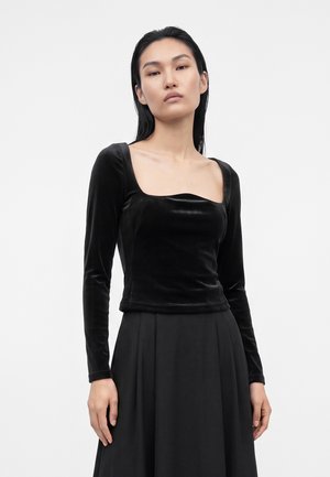 Femme portant un haut en velours noir à manches longues avec un décolleté carré et une jupe plissée noire, se tenant devant un fond clair uni.