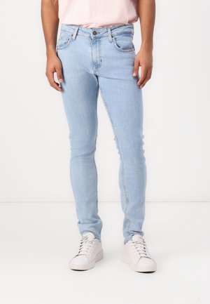 Person, der hellblaue Slim-Fit-Jeans, weiße Turnschuhe und ein in die Hose gestecktes, hellrosa Hemd trägt, steht vor einem schlichten weißen Hintergrund.