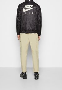 Mustat Nike-takit, joissa on valkoinen "Nike Air" -logo, rakenteellinen kangas, vetoketjukaulus, yhdistettynä beigeihin cargo-housuihin ja mustiin lenkkareihin.