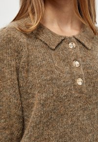Žena v hnědém chlupatém pleteném svetru s širokým límcem a třemi mramorovými knoflíky vpředu.