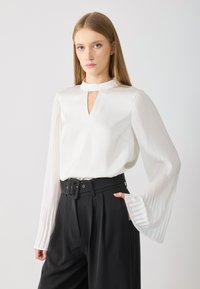 White blouse with a keyhole neckline and pleated long sleeves, paired with high-waisted black trousers and a wide belt.