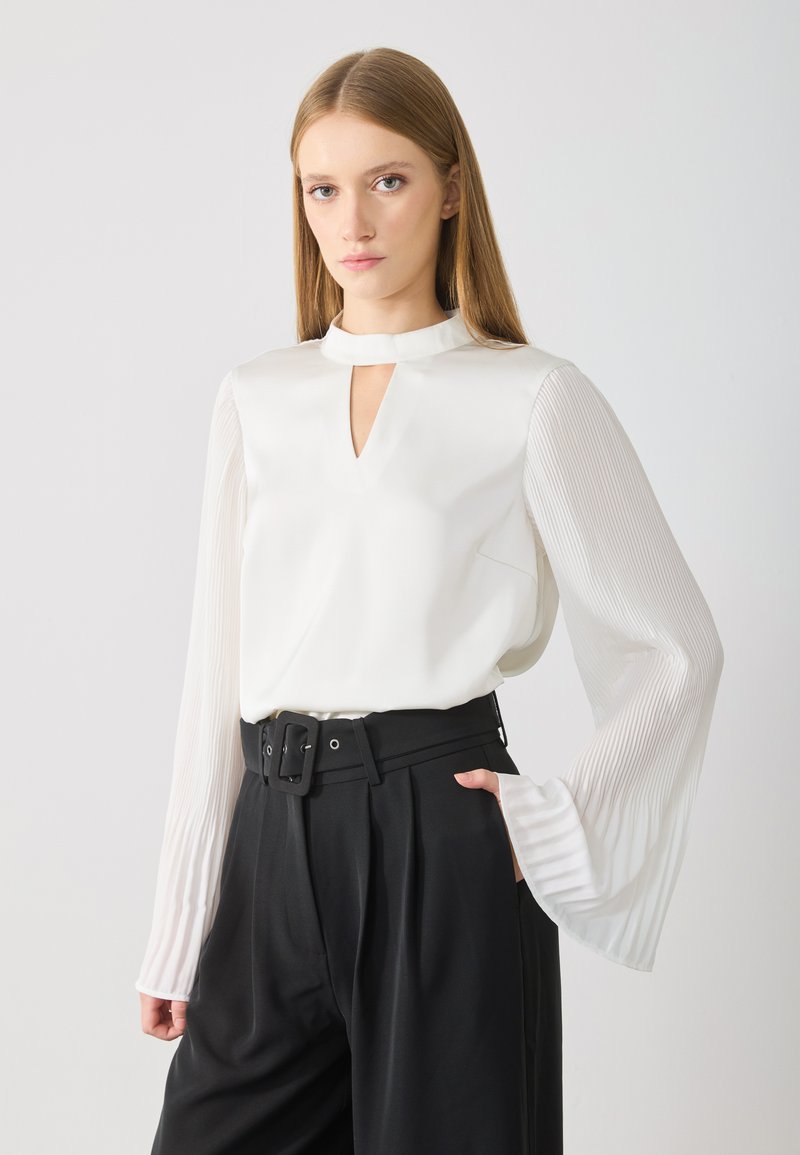 White blouse with a keyhole neckline and pleated long sleeves, paired with high-waisted black trousers and a wide belt.