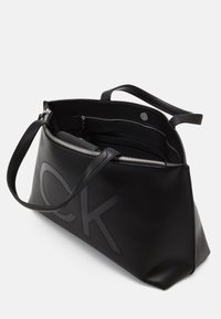 Sac à main en cuir noir avec un logo CK bien en vue, une fermeture éclair et deux poignées. Texture lisse avec un intérieur spacieux et des poches intérieures.