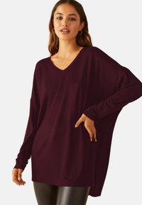 Burgundská blúzka s dlhým rukávom a výstrihom do V s voľným, oversized strihom. Mäkka textúra, spustené ramená a bočné rozparky pre väčšiu pohyblivosť a pohodlie.