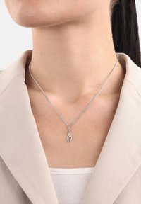 Collana con pendente a forma di chiave di violino argentata indossata al collo con giacca beige e top bianco.