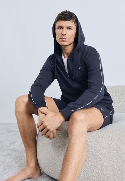 Marineblå hættetrøje med lynlås, frontlommer og hvidt logo på brystet, kombineret med matchende shorts. Blødt stof, afslappet pasform, placeret på en tekstureret overflade.