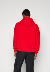 Felpa rossa con cappuccio grande, polsini elastici e orlo. Lo sfondo è semplice, mettendo in evidenza la texture e il colore vivace del capo.
