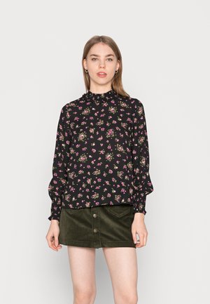 Blusa preta floral com colarinho e mangas com babados, apresentando pequenas flores rosas e verdes. Usada com uma saia de cotim verde escuro com botões.
